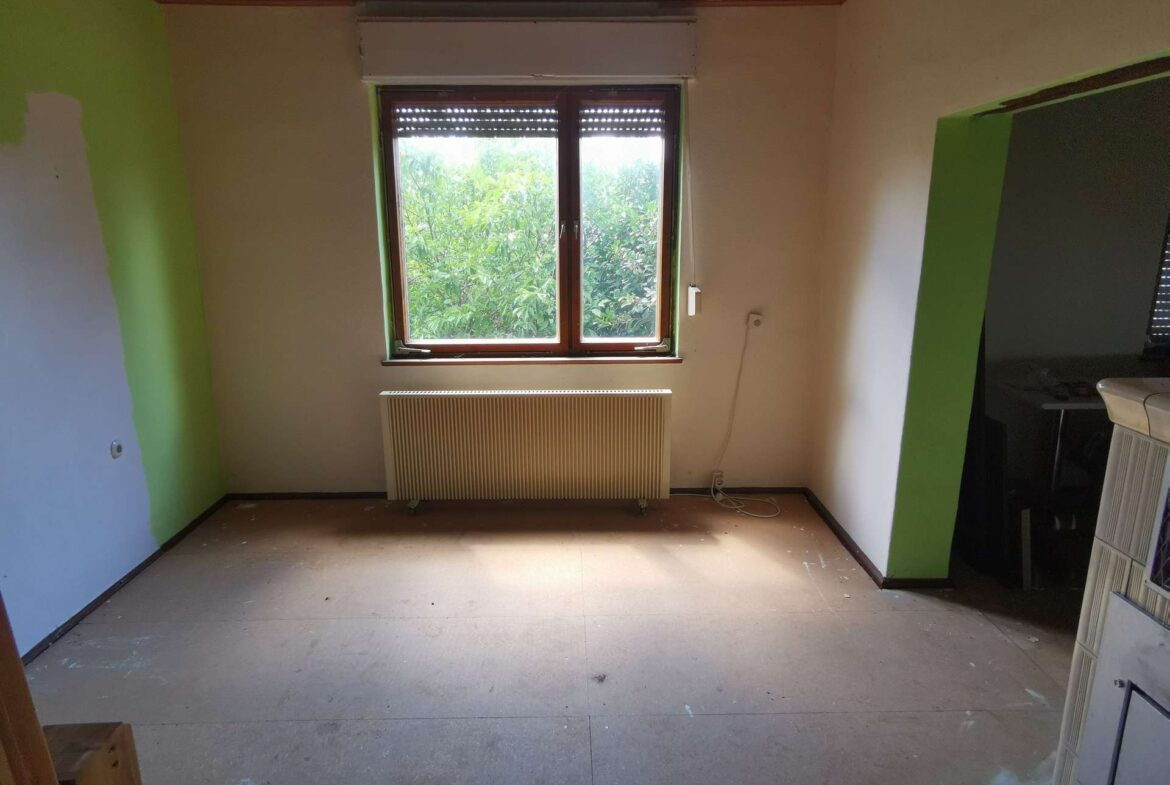 Empty room under renovation with a large window looking out to green trees, and a radiator beneath the window; lime-green wall on the left.