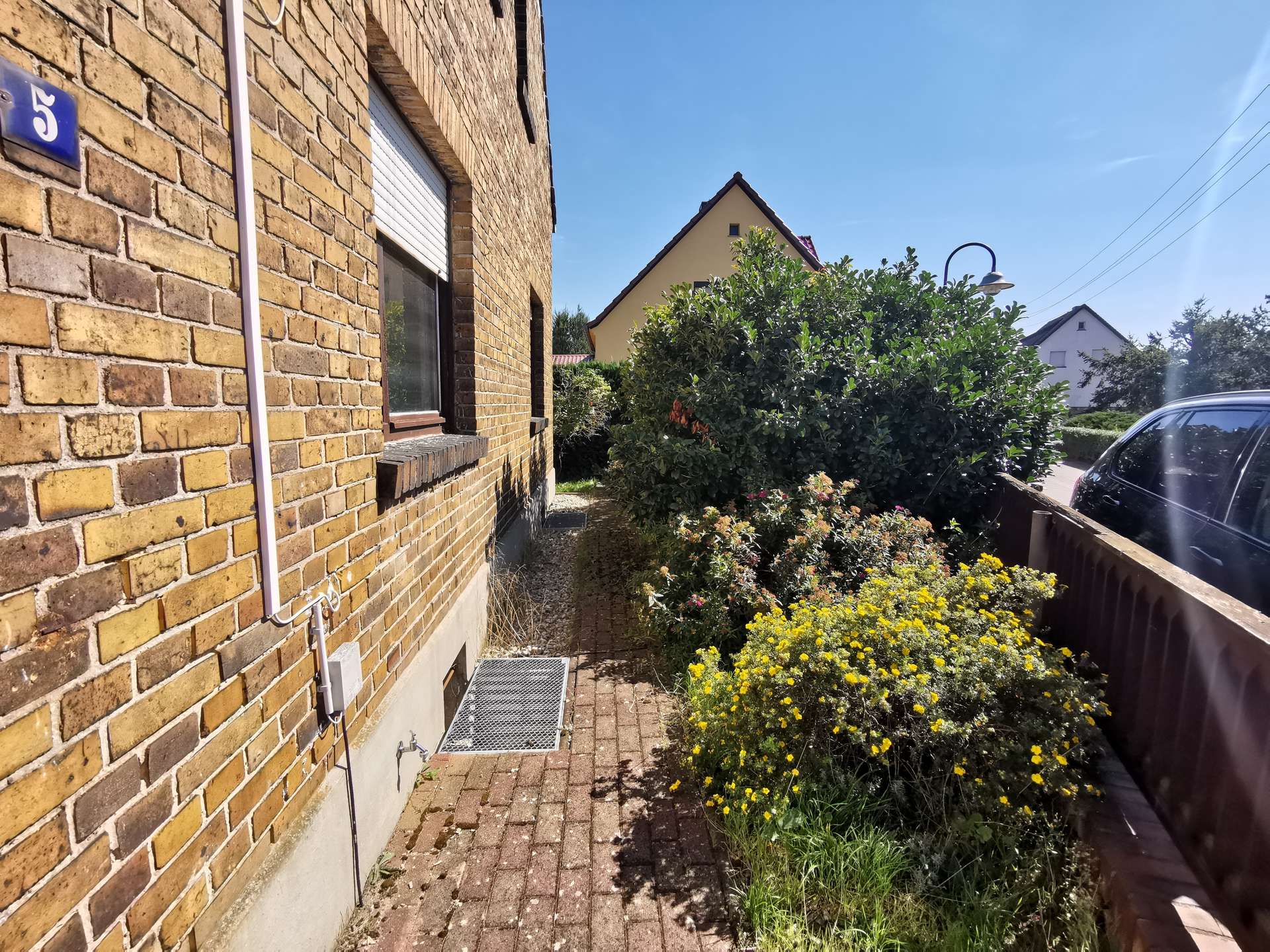 Brick house wall with a metal drainpipe, window, and a small metal grate on the ground beside a paved walkway, sunny day.