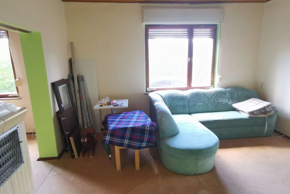 Living room with a light green sectional sofa near a window, cluttered by a small table, plaid chair cover, and stacked frames/ladder pieces against a lime column.
