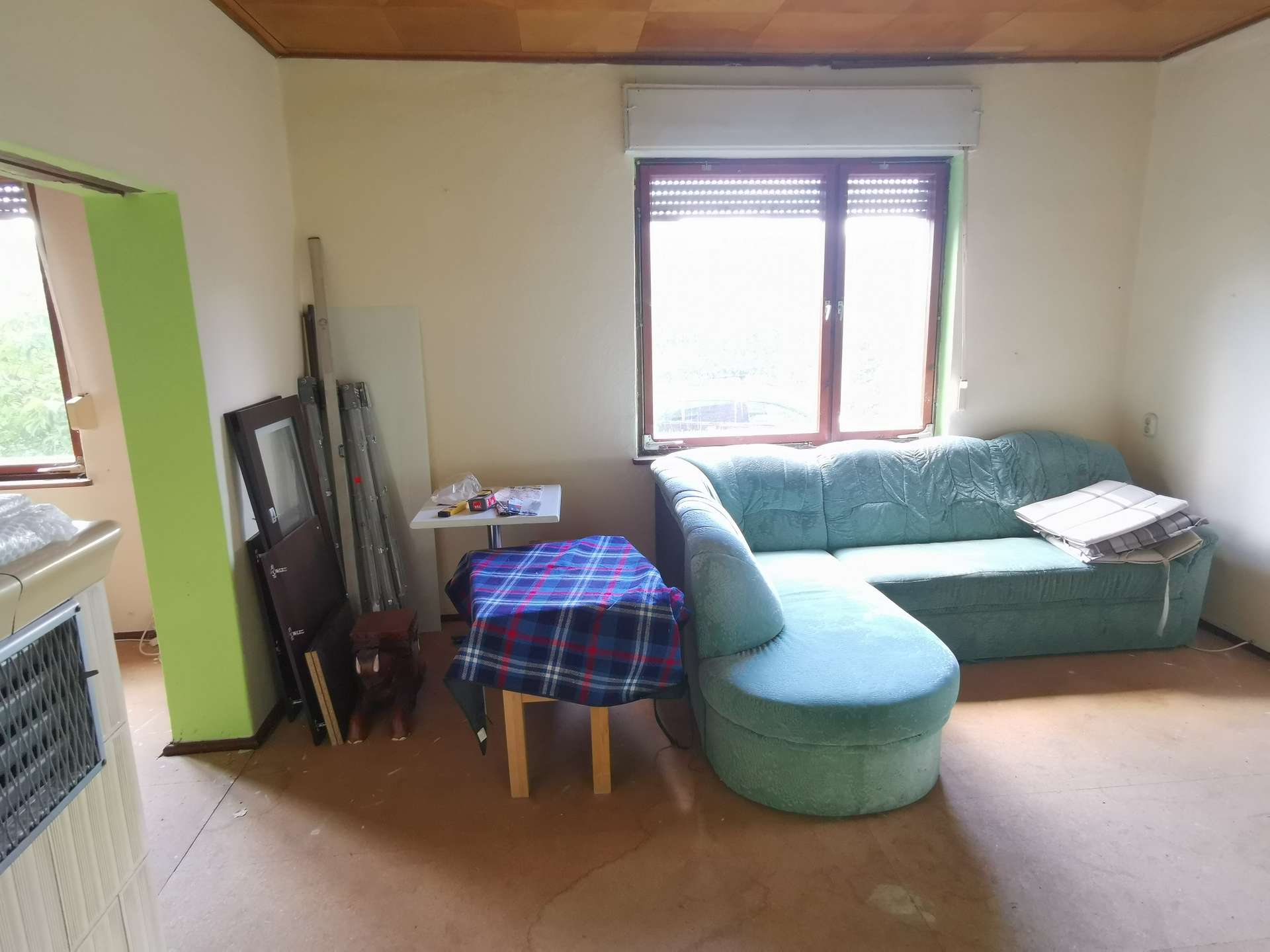 Living room with a light green sectional sofa near a window, cluttered by a small table, plaid chair cover, and stacked frames/ladder pieces against a lime column.