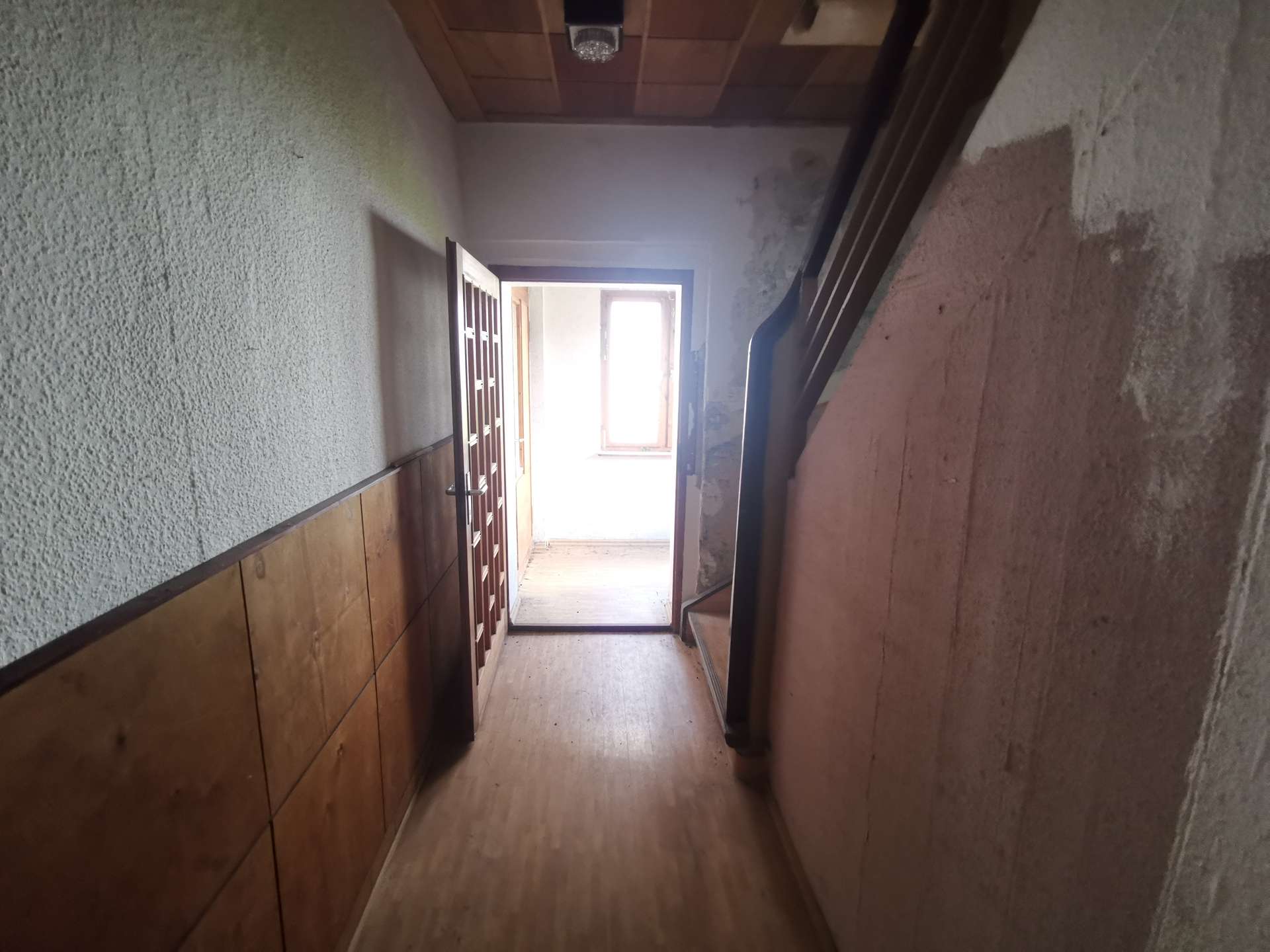 Narrow hallway with wood paneling on the left, a staircase on the right, and an open door at the far end letting in light.