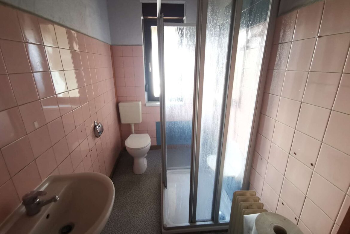 Small bathroom with pink tiled walls, a white toilet and sink, and a glass-enclosed corner shower with sliding doors, plus a window and towel rack visible.