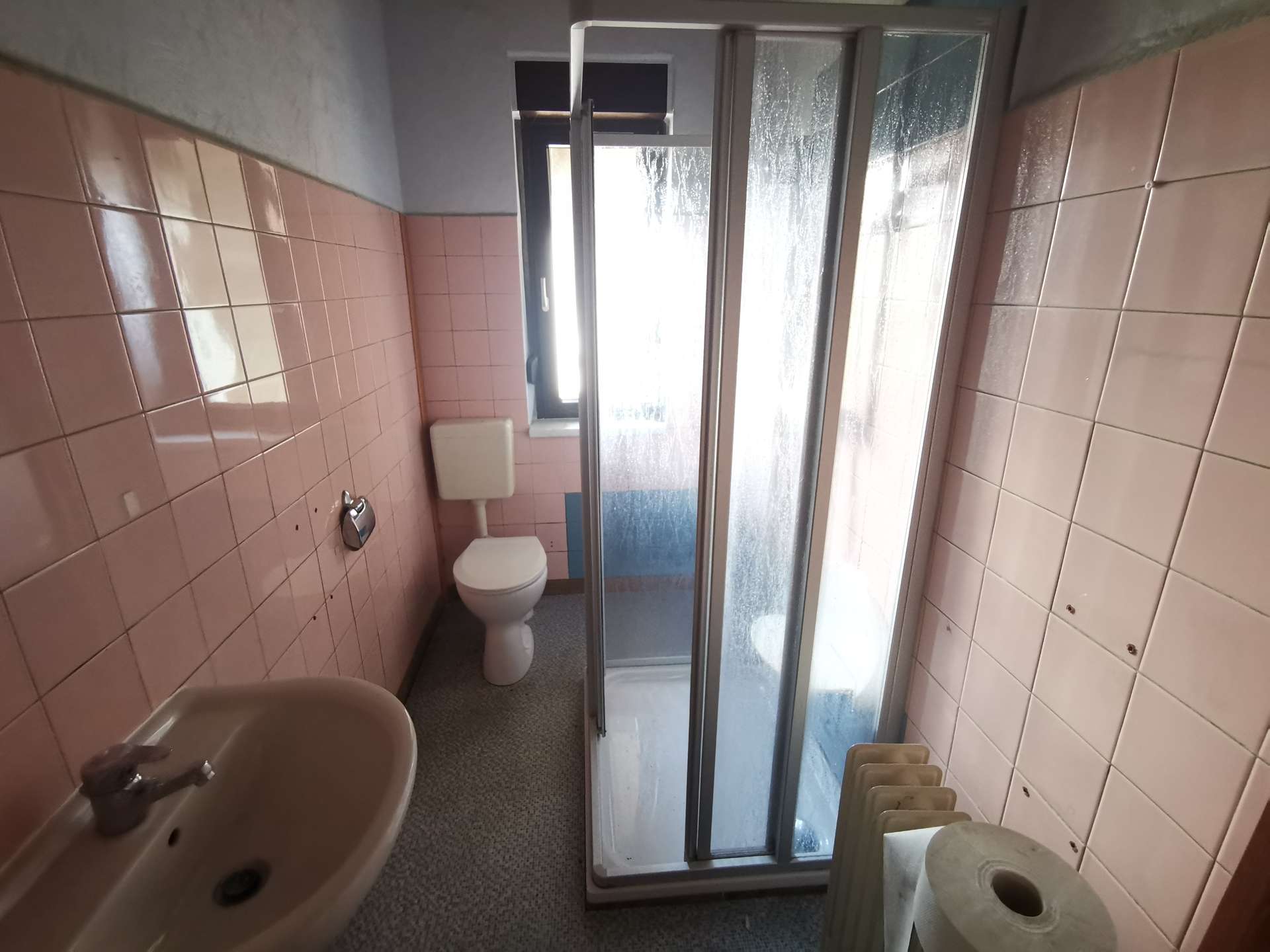 Small bathroom with pink tiled walls, a white toilet and sink, and a glass-enclosed corner shower with sliding doors, plus a window and towel rack visible.