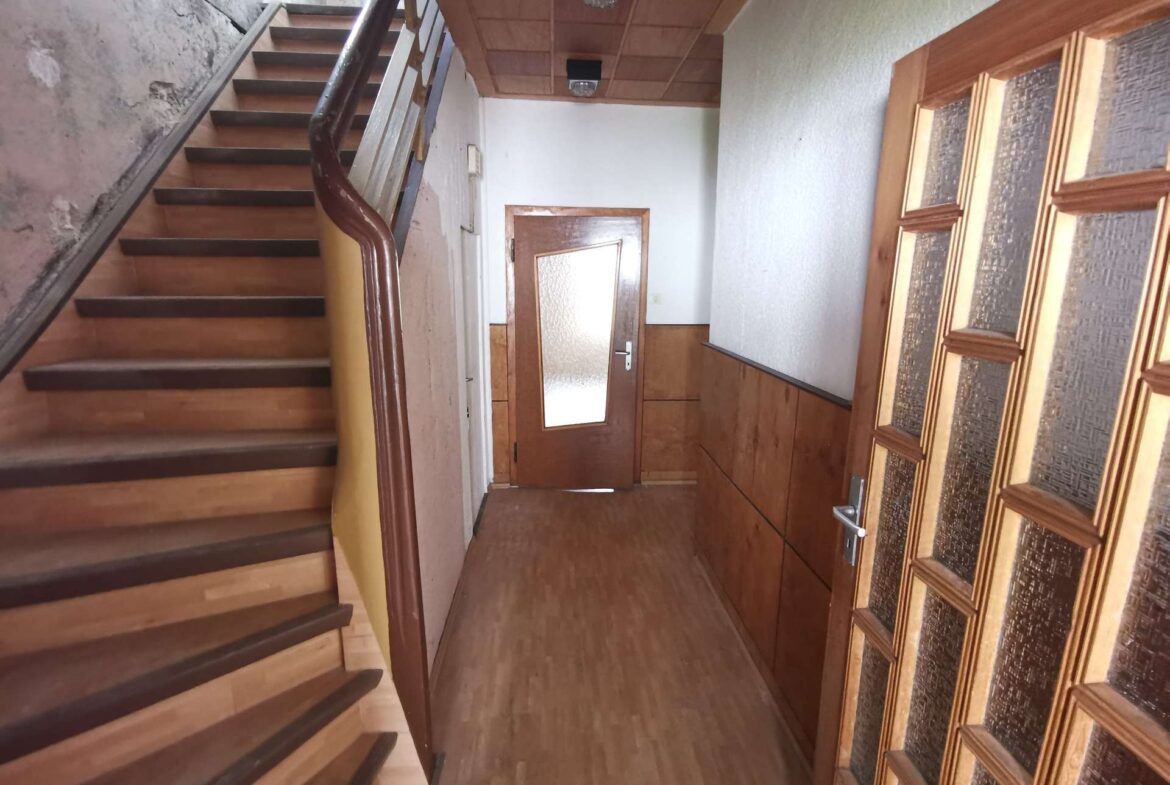 Narrow indoor stairwell with wooden steps and dark handrail leading to a wooden door at the end of a dim hallway.