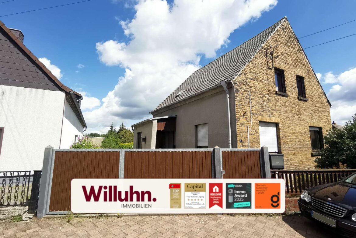 Front view of a brick house with a steep gabled roof behind a brown wooden fence, under a blue sky with clouds; a real estate sign in the foreground reading 'Willuhn Immobilien'.