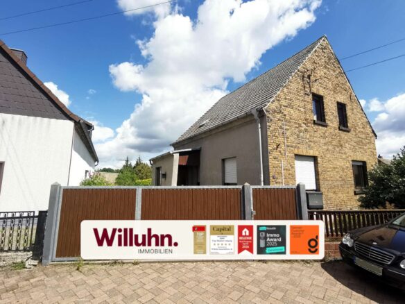 Front view of a brick house with a steep gabled roof behind a brown wooden fence, under a blue sky with clouds; a real estate sign in the foreground reading 'Willuhn Immobilien'.