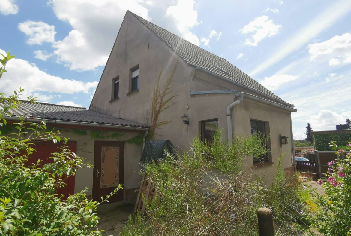 Two-story beige house with a steep, tiled roof and vines climbing the wall, set against a blue sky with clouds.