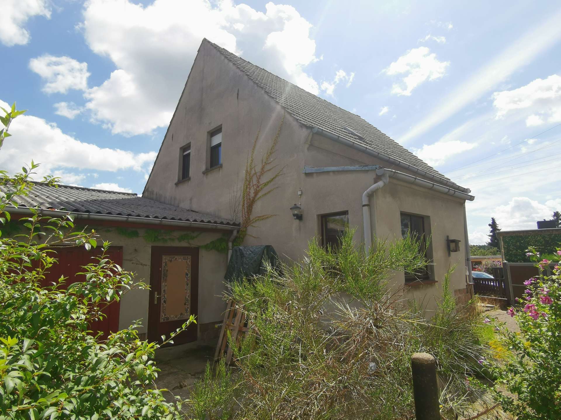 Two-story beige house with a steep, tiled roof and vines climbing the wall, set against a blue sky with clouds.