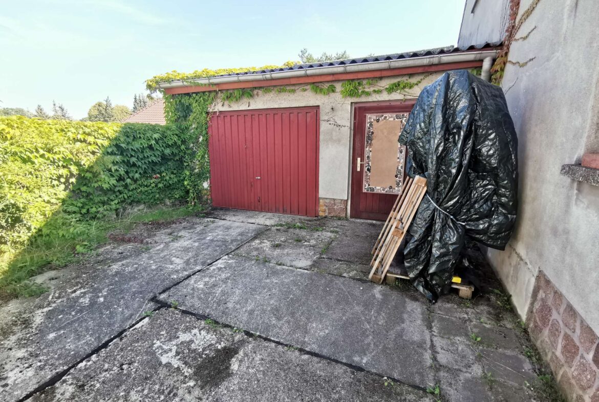 Outdoor scene of a building with red garage doors, ivy climbing the wall, and a large black tarp covering items with wooden pallets nearby.