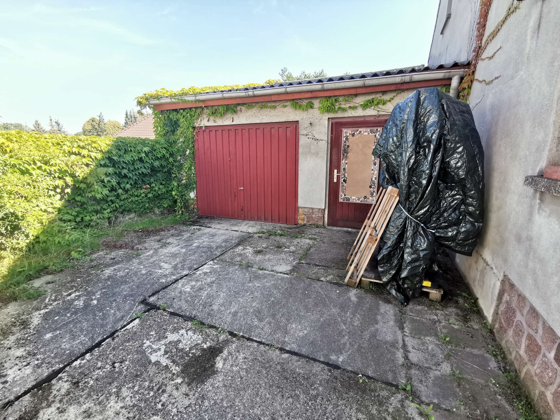 Outdoor scene of a building with red garage doors, ivy climbing the wall, and a large black tarp covering items with wooden pallets nearby.