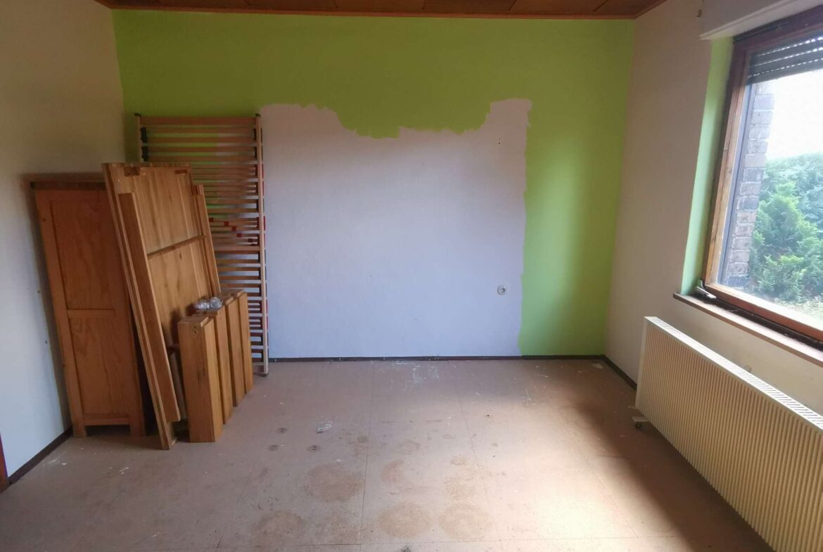 A sparsely furnished room being repainted: a bright green wall with a large white patch, and wooden furniture stacked along the left wall beside a ladder-like bed frame. A radiator runs along the right wall beneath a large window.
