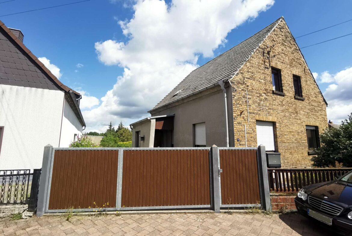 Brick house with a steep triangular gable roof, behind a tall brown wooden fence, under a blue sky with white clouds.