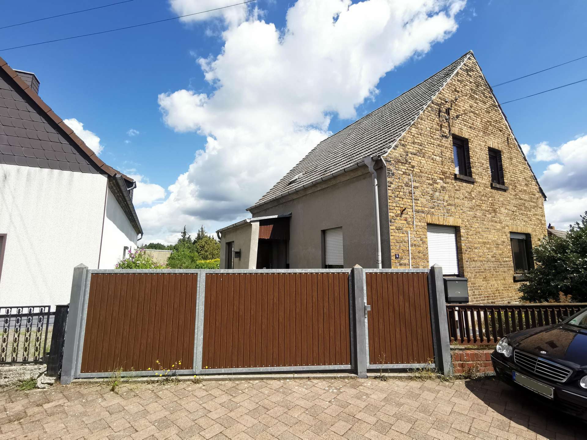 Brick house with a steep triangular gable roof, behind a tall brown wooden fence, under a blue sky with white clouds.