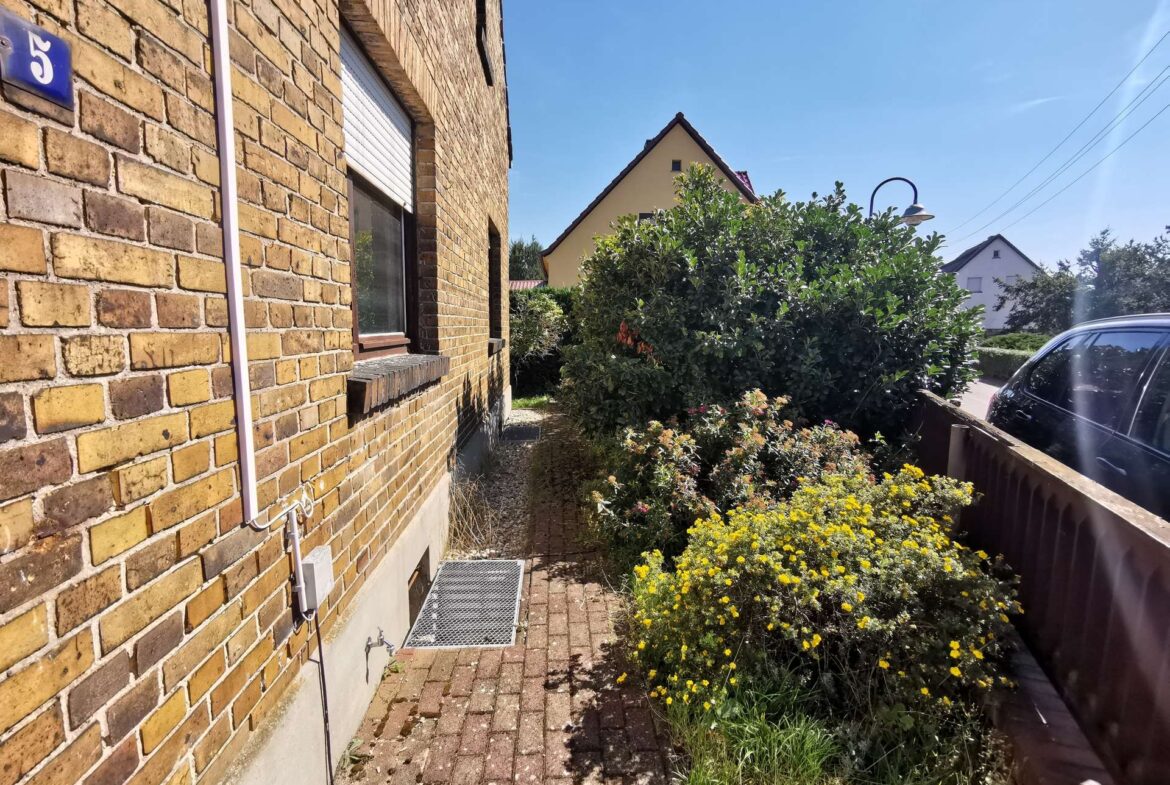 Brick house exterior with a window and downspout, a metal grate on the path, and a garden bed of yellow flowers under a blue sky, with a car on the right.