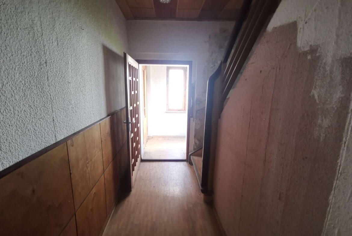 Narrow indoor hallway with wooden paneling on the left, stairs on the right, and a bright doorway at the end letting in outside light.