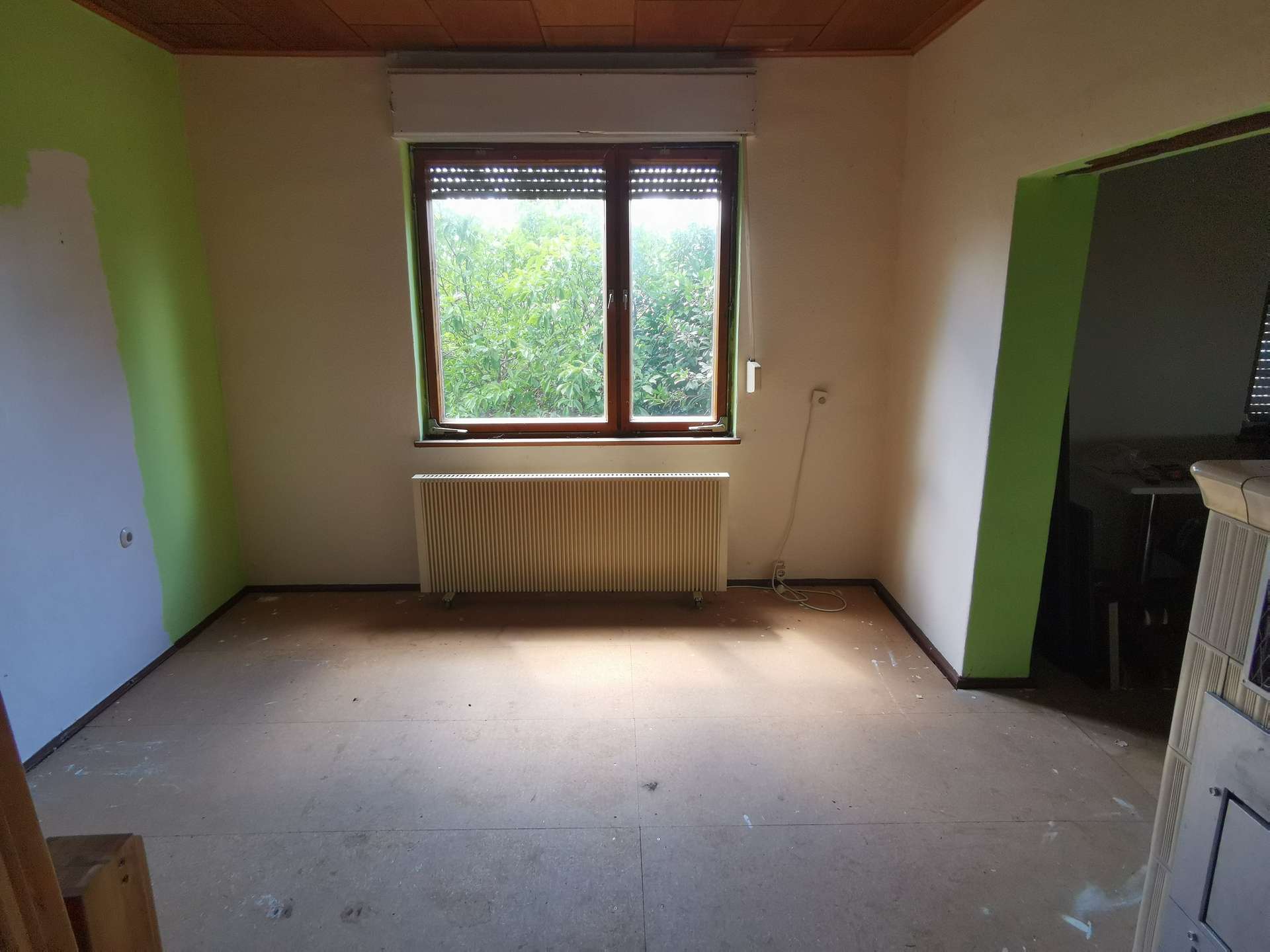 Empty room under renovation with a large central window, beige walls, and a green accent wall on the left; a radiator below the window and a dark cabinet or wardrobe on the right.