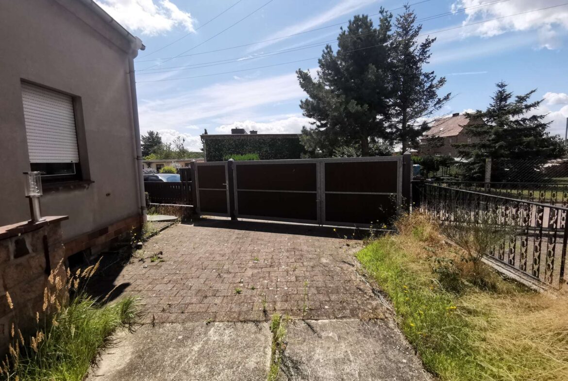 Driveway in front of a beige house with a closed window shutter, a metal gate, and overgrown grass under a blue sky.