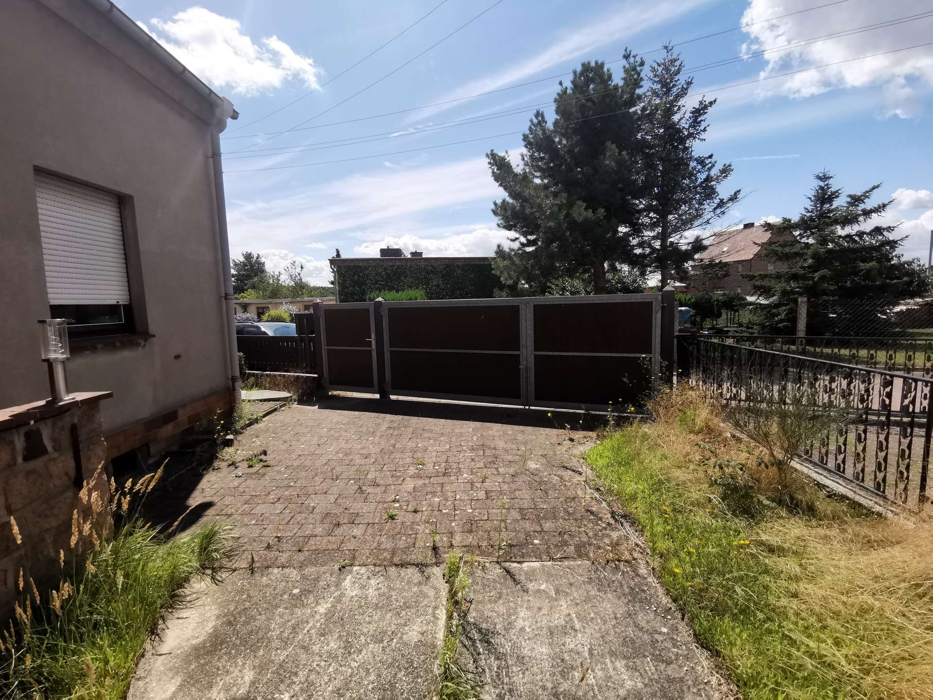 Driveway in front of a beige house with a closed window shutter, a metal gate, and overgrown grass under a blue sky.