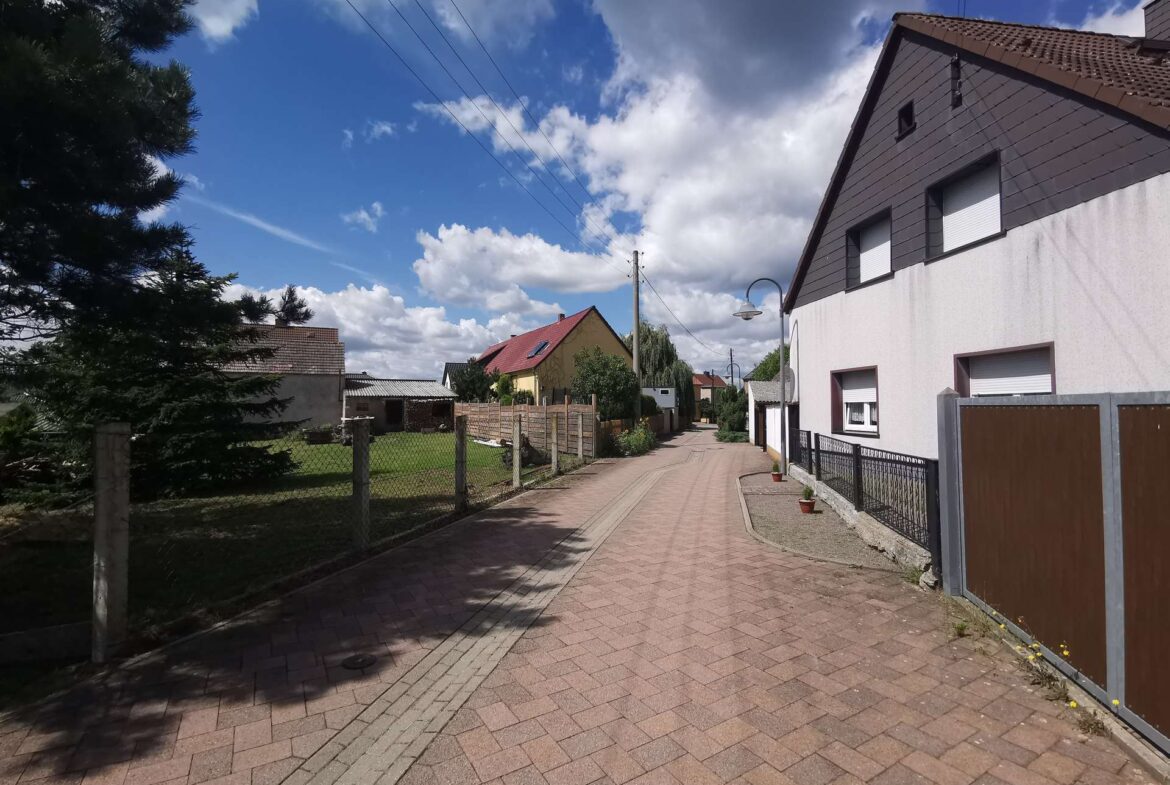 Residential street scene with brick-paved sidewalk, fences, and row of houses under a blue, partly cloudy sky.