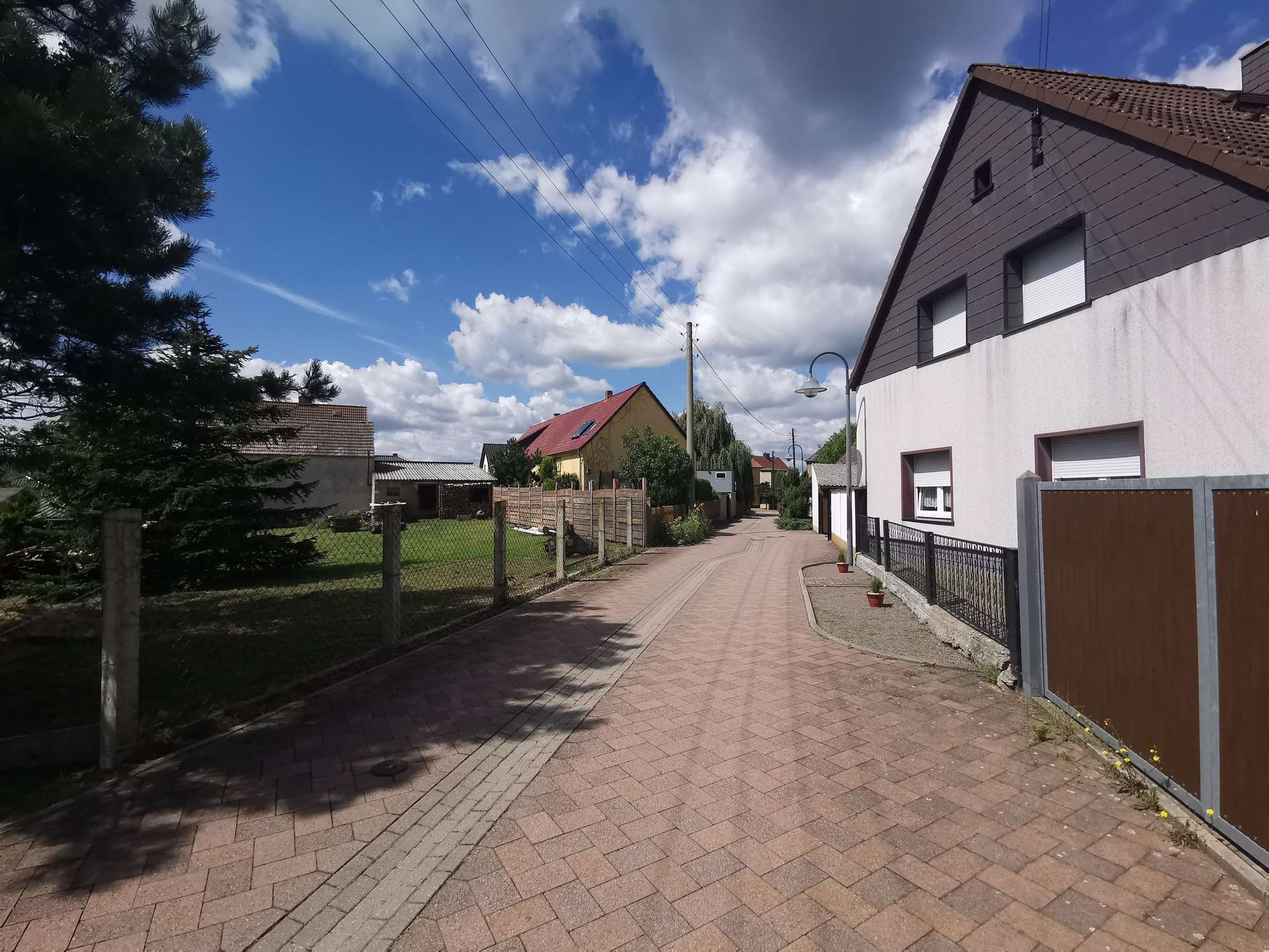Residential street scene with brick-paved sidewalk, fences, and row of houses under a blue, partly cloudy sky.