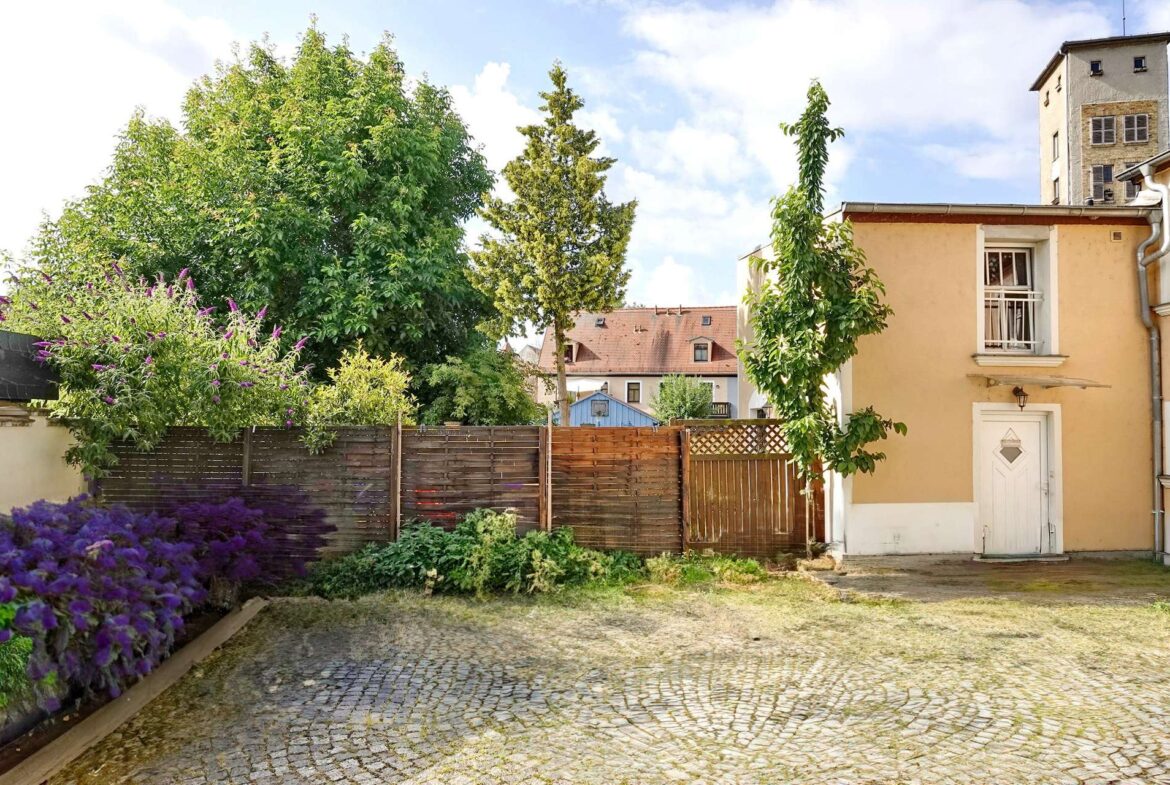 A sunny courtyard with a cobblestone path, wooden fence, and colorful flowering bushes beside a peach-colored building.