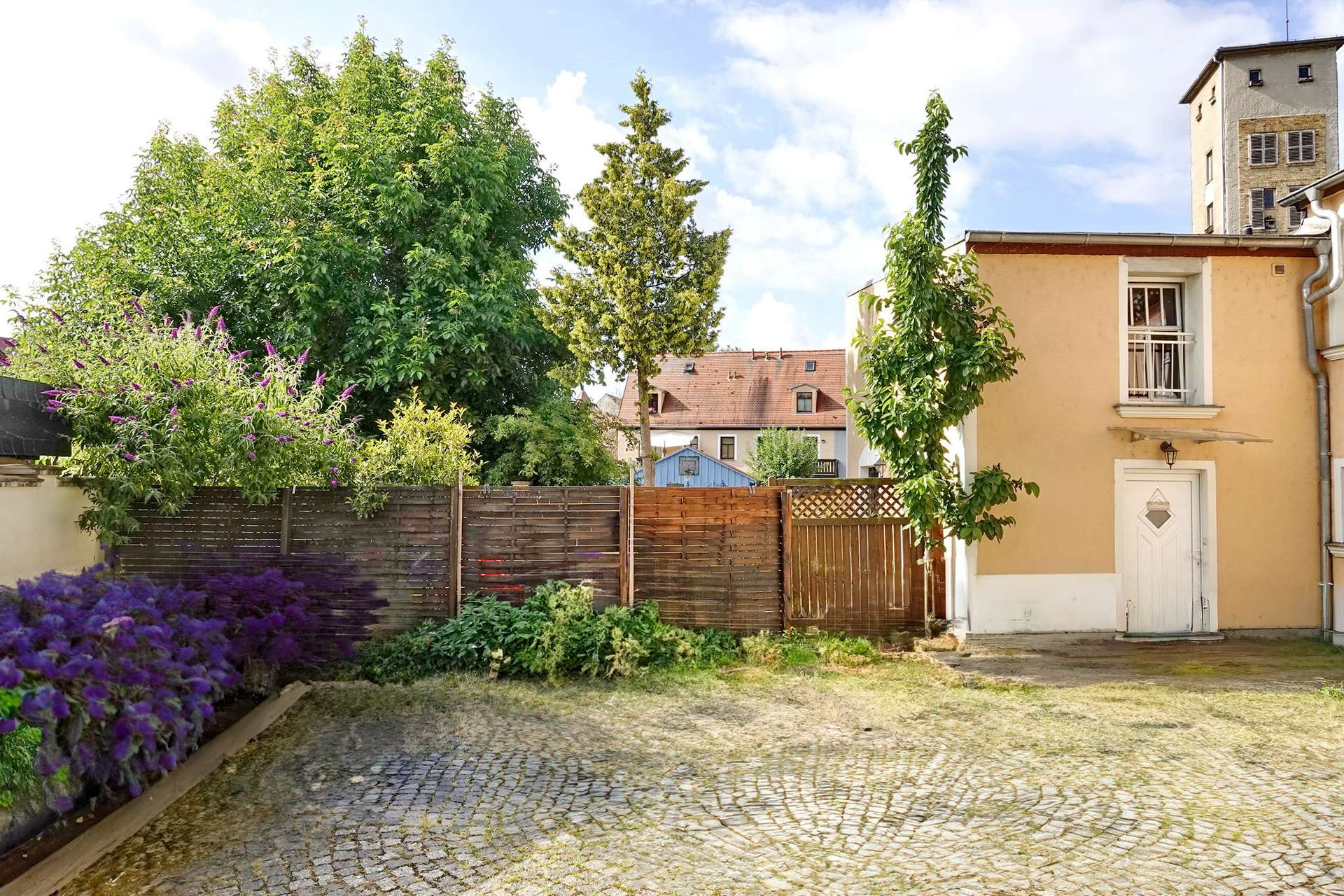 A sunny courtyard with a cobblestone path, wooden fence, and colorful flowering bushes beside a peach-colored building.