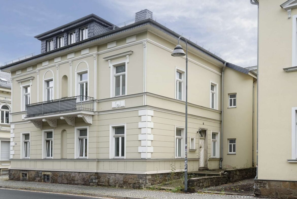Three-story beige building with white decorative trim, arched second‑floor windows, and a black wrought‑iron balcony along a cobblestone street.