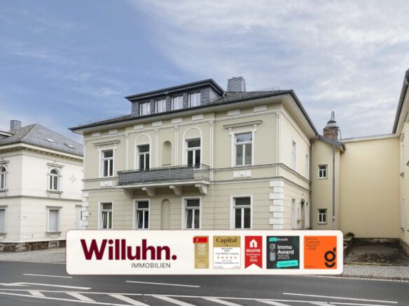 Beige, multi-story building on a city street with a large Willuhn Immobilien real estate sign and award stickers in front.