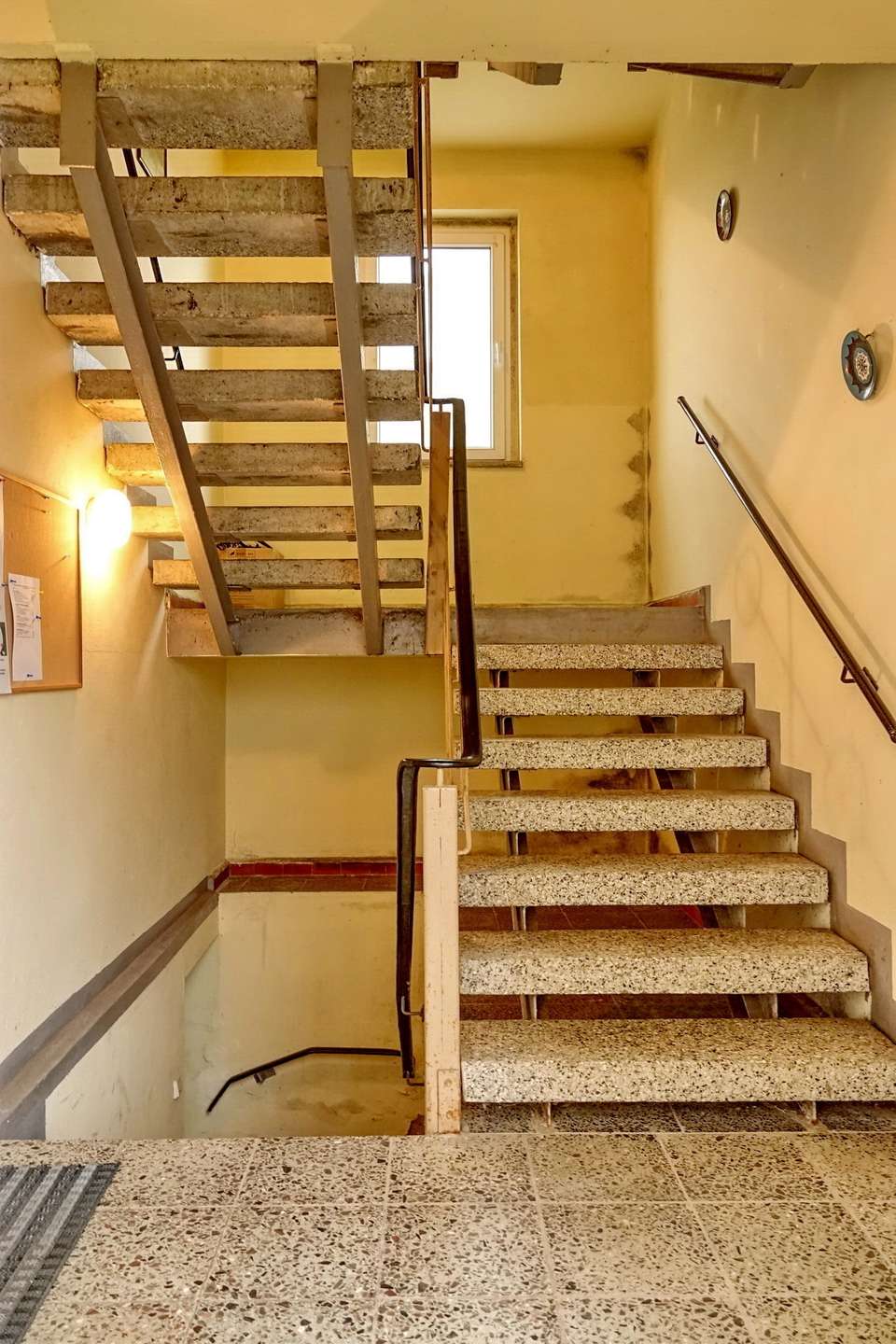 Concrete stairwell with yellow walls, metal railings, and a window at the landing—industrial building interior