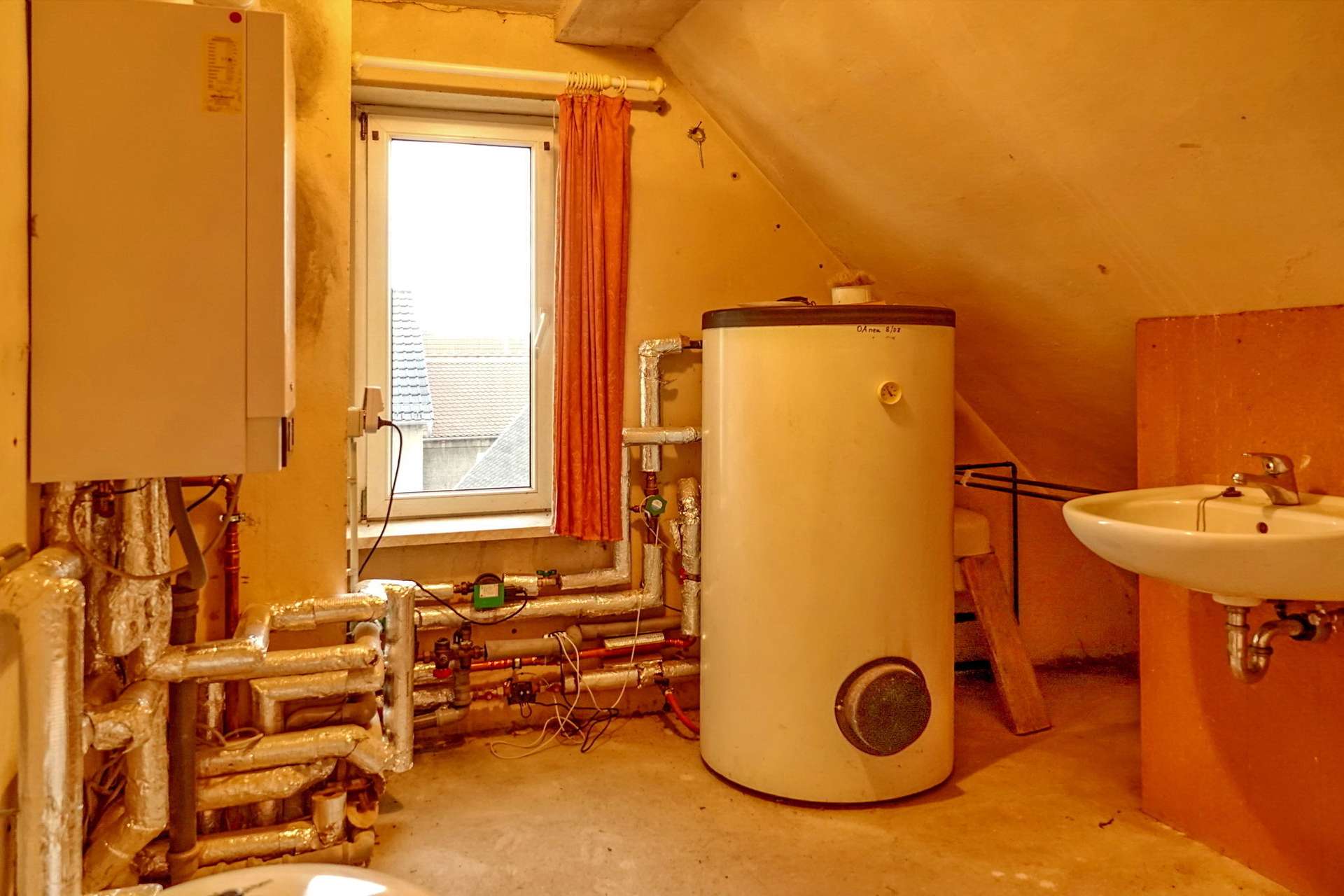 Basement utility room with a tall water heater, exposed pipes, and a window with an orange curtain beside a sink.