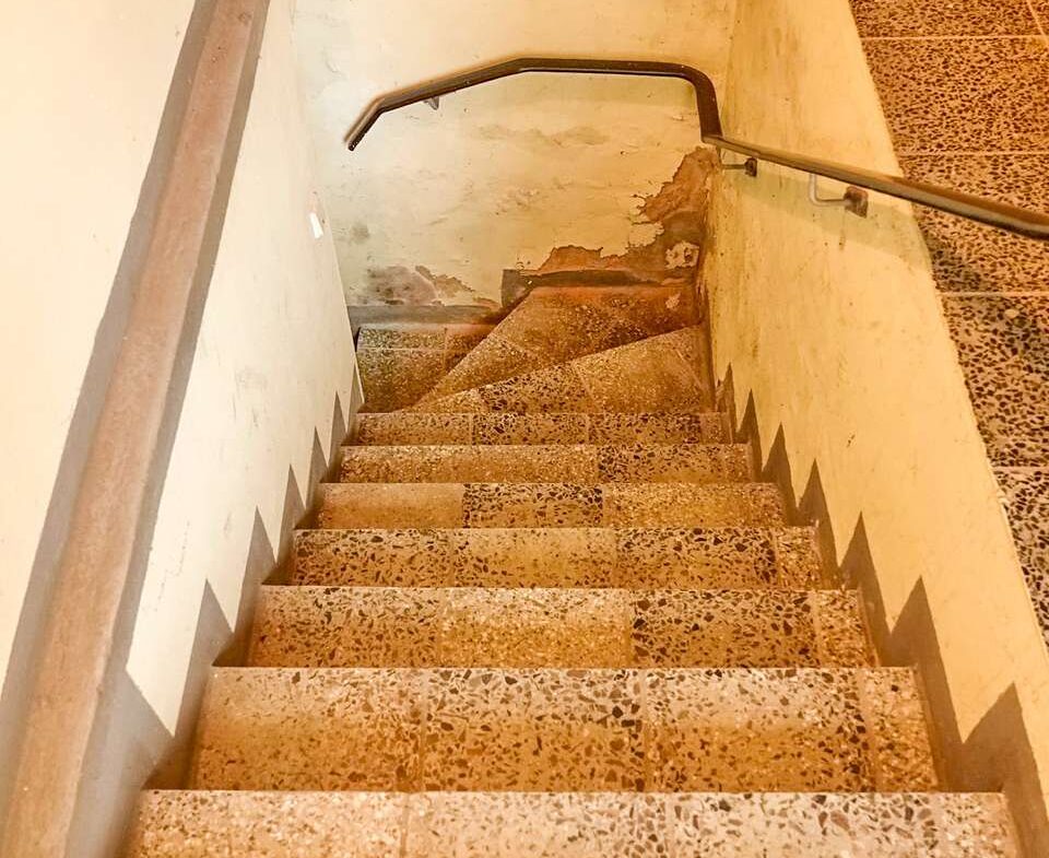 Indoor stairwell with terrazzo steps, metal handrail along the right, and worn, peeling walls at the bottom.