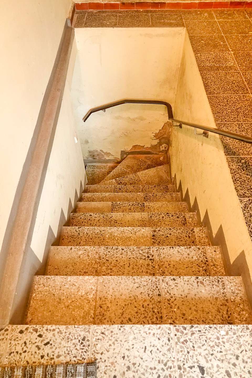 Indoor stairwell with terrazzo steps, metal handrail along the right, and worn, peeling walls at the bottom.