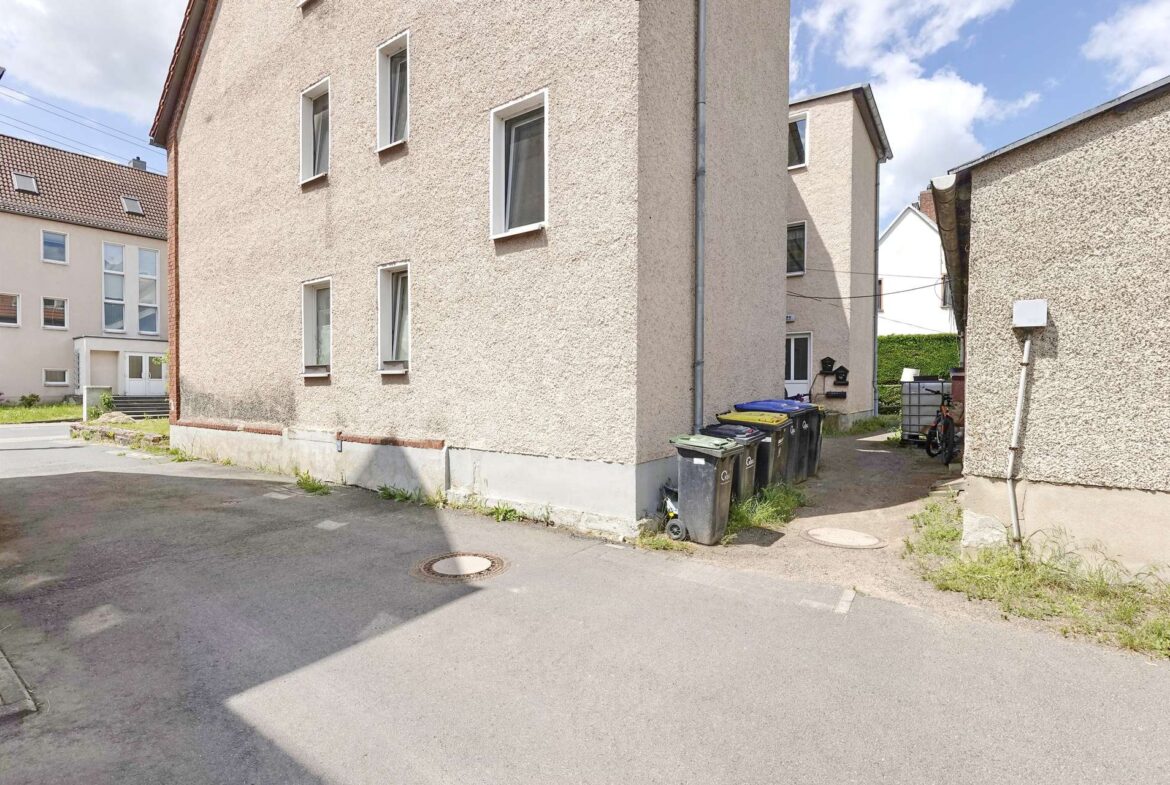 Beige stucco apartment building along a narrow alley with a row of waste bins and a parked bicycle nearby