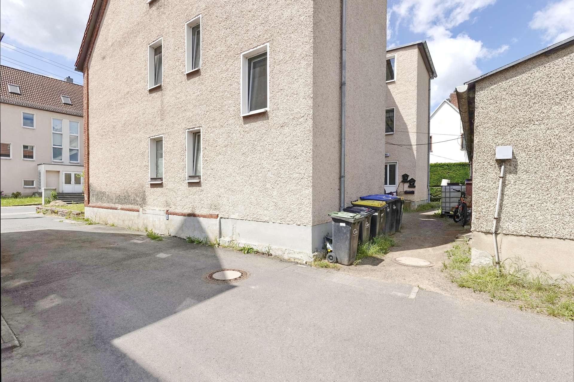 Beige stucco apartment building along a narrow alley with a row of waste bins and a parked bicycle nearby