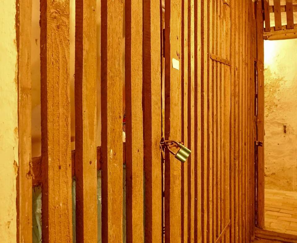 Narrow hallway with vertical wooden slats forming a cell-like barrier and a green padlock on a door handle in warm yellow light
