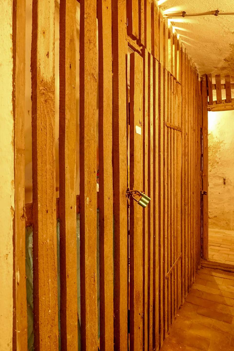 Narrow hallway with vertical wooden slats forming a cell-like barrier and a green padlock on a door handle in warm yellow light