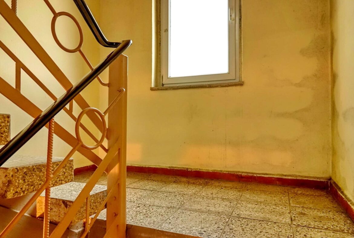 Interior stairwell with orange metal railing and terrazzo steps, yellow walls, and a bright window illuminating the space.