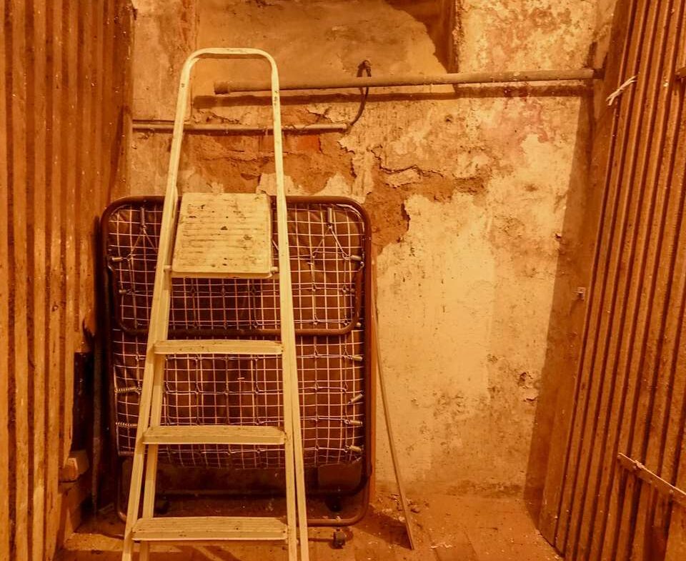 Storage area with a white step ladder leaning against a folded metal bed frame and rough peach walls under a small high window.