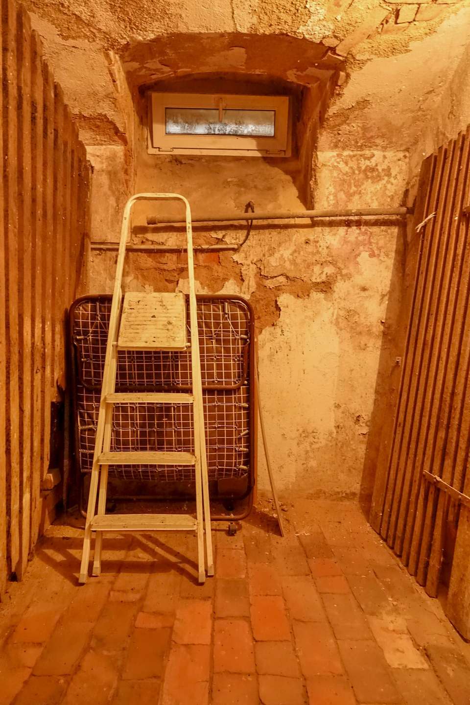 Storage area with a white step ladder leaning against a folded metal bed frame and rough peach walls under a small high window.