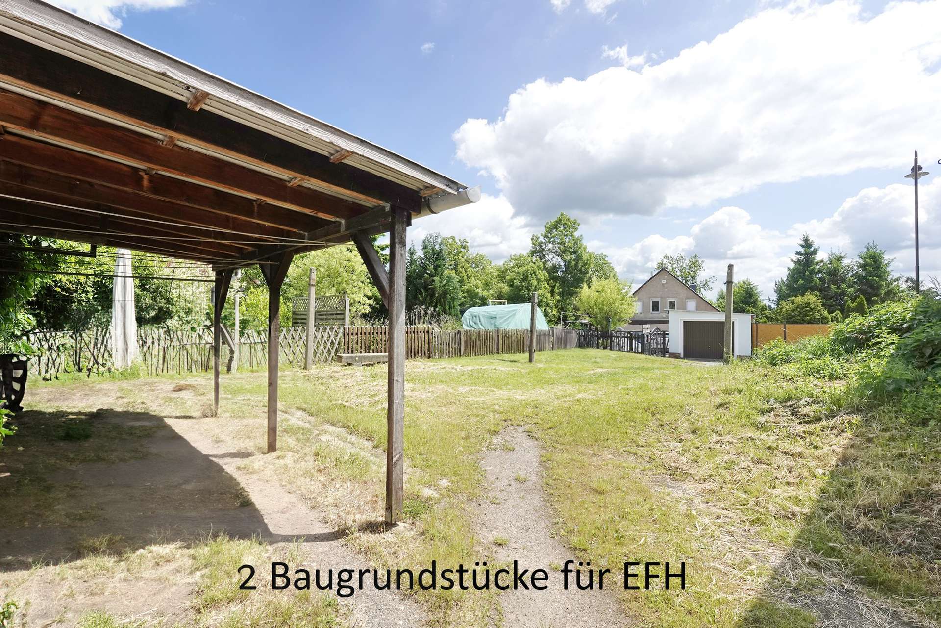Backyard scene with a wooden carport on the left, a grassy area and fence, and a house in the distance; overlay text advertises two building plots for single-family homes.