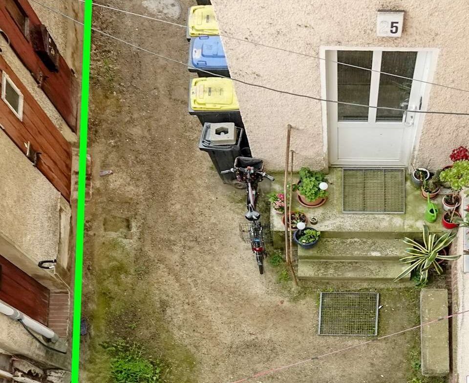 Bird's-eye view of a narrow courtyard between buildings, with a row of waste bins, a bicycle, and potted plants near a beige door; a green outline marks a rectangular area.