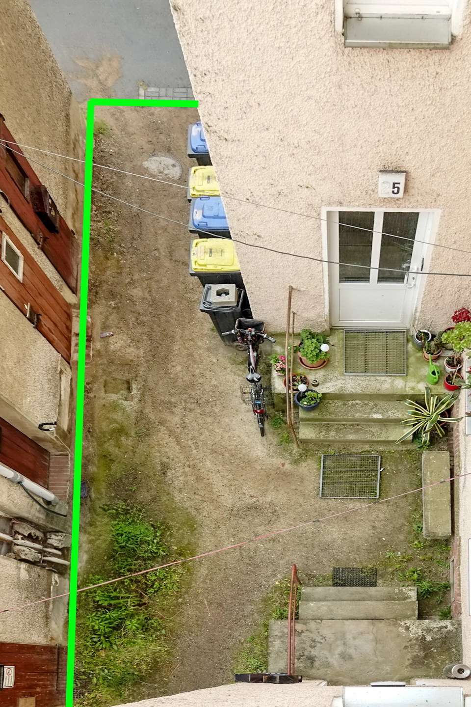 Bird's-eye view of a narrow courtyard between buildings, with a row of waste bins, a bicycle, and potted plants near a beige door; a green outline marks a rectangular area.