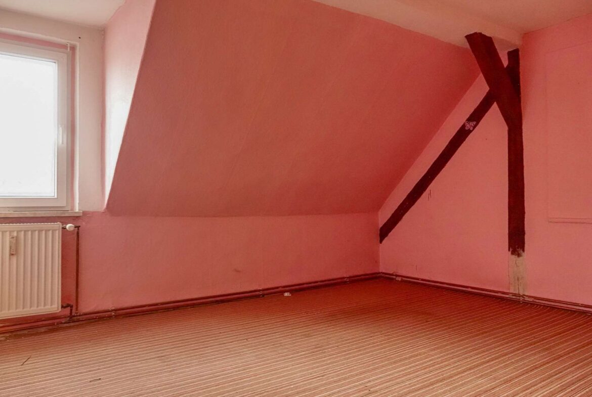 Empty attic room with pink walls, a sloped ceiling, and a dark diagonal support beam near the corner, window on the left, radiator below it.