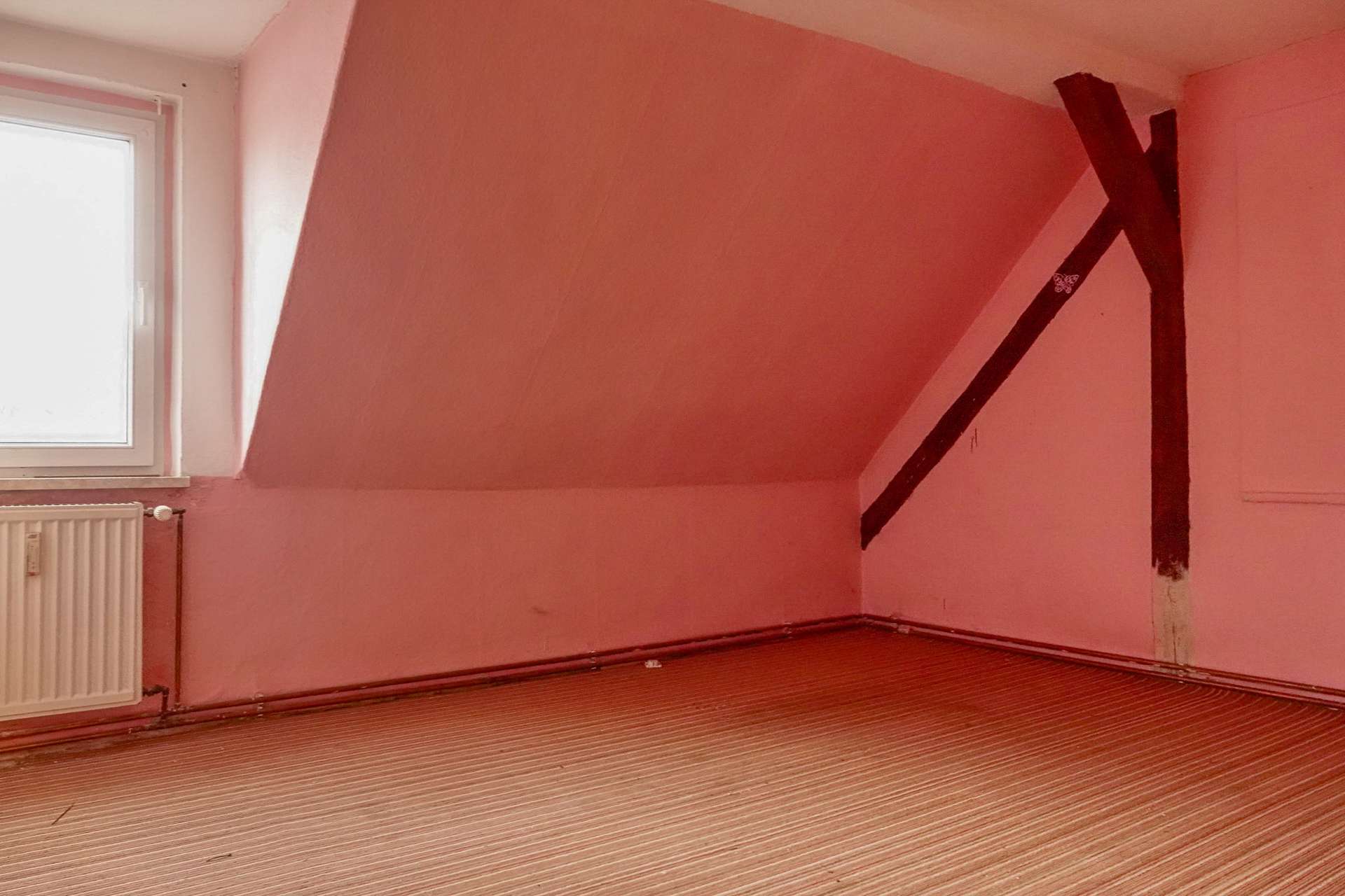 Empty attic room with pink walls, a sloped ceiling, and a dark diagonal support beam near the corner, window on the left, radiator below it.