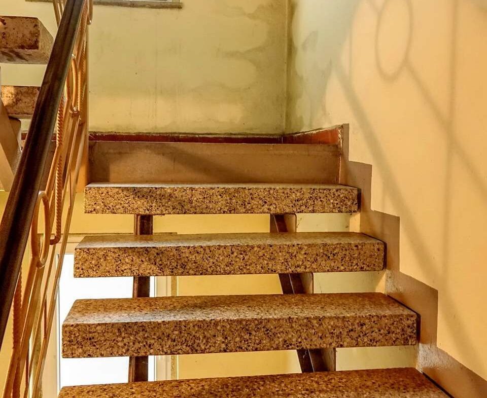 Interior stairwell with exposed terrazzo steps and a metal handrail along the left, yellow walls, and graffiti on the right wall near a window overhead
