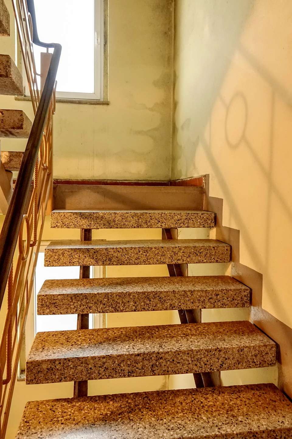 Interior stairwell with exposed terrazzo steps and a metal handrail along the left, yellow walls, and graffiti on the right wall near a window overhead