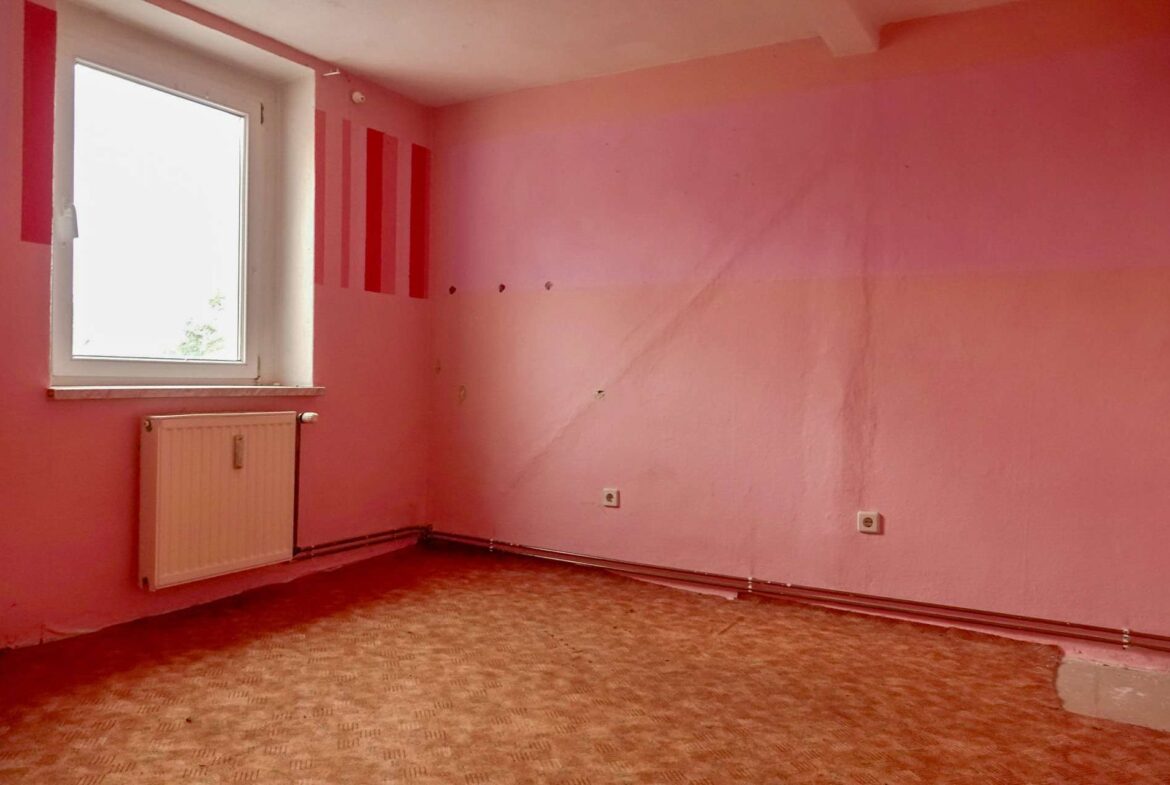 Empty pink-walled room with a white window, radiator beneath, and worn patterned carpet; left wall has red vertical stripes and small holes.