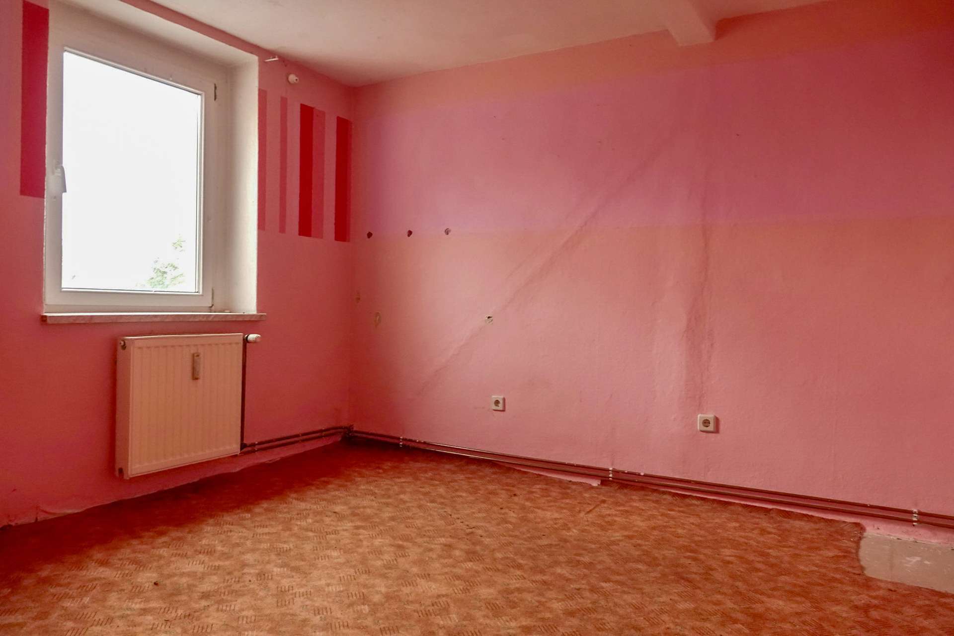 Empty pink-walled room with a white window, radiator beneath, and worn patterned carpet; left wall has red vertical stripes and small holes.