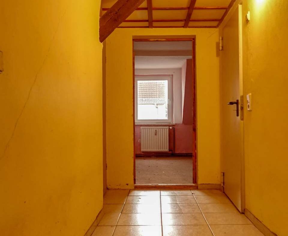 Yellow hallway with tiled floor, exposed wooden ceiling beams, and a doorway revealing a sunlit room with a radiator and window at the end.