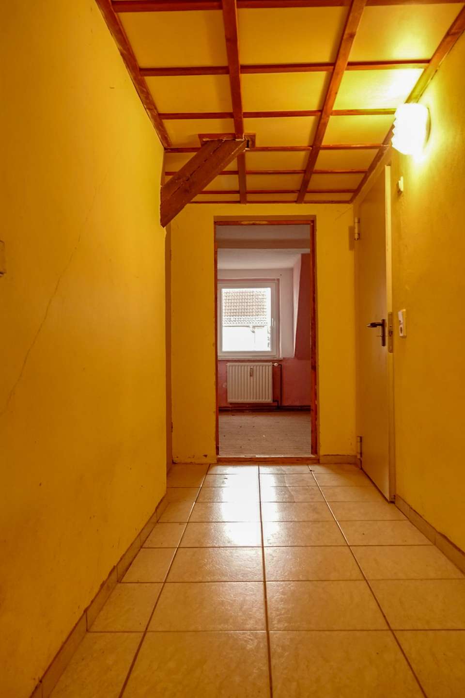 Yellow hallway with tiled floor, exposed wooden ceiling beams, and a doorway revealing a sunlit room with a radiator and window at the end.